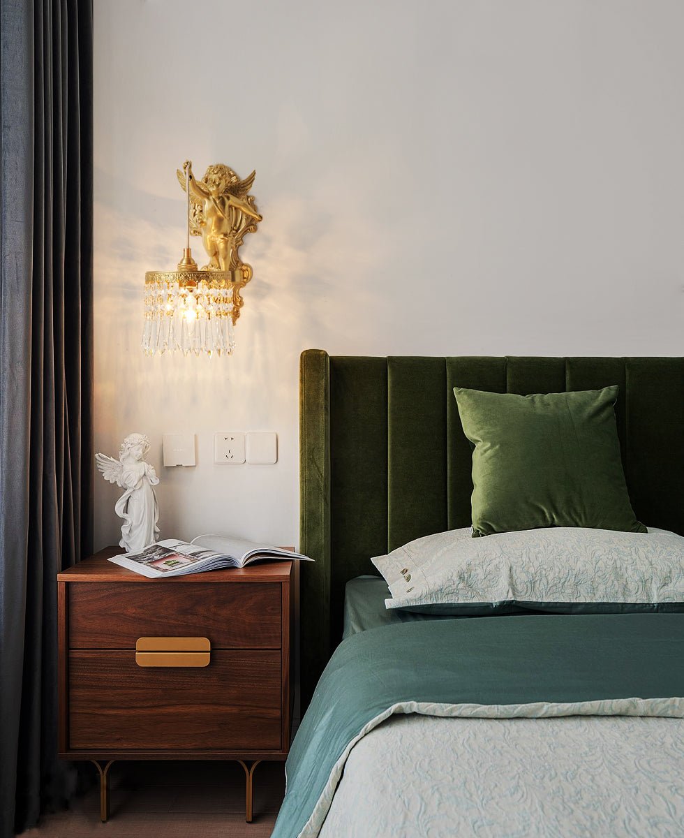 Above a walnut bedside table, a Clérachic cherub wall light sits on a pale wall beside a green velvet headboard.