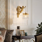 A Clérachic cherub wall light with a crystal-fringed shade on a panelled wall above a side table with framed photographs.
