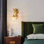 Close to a green velvet headboard, a Clérachic cherub wall light glows above a walnut bedside table with an open magazine.
