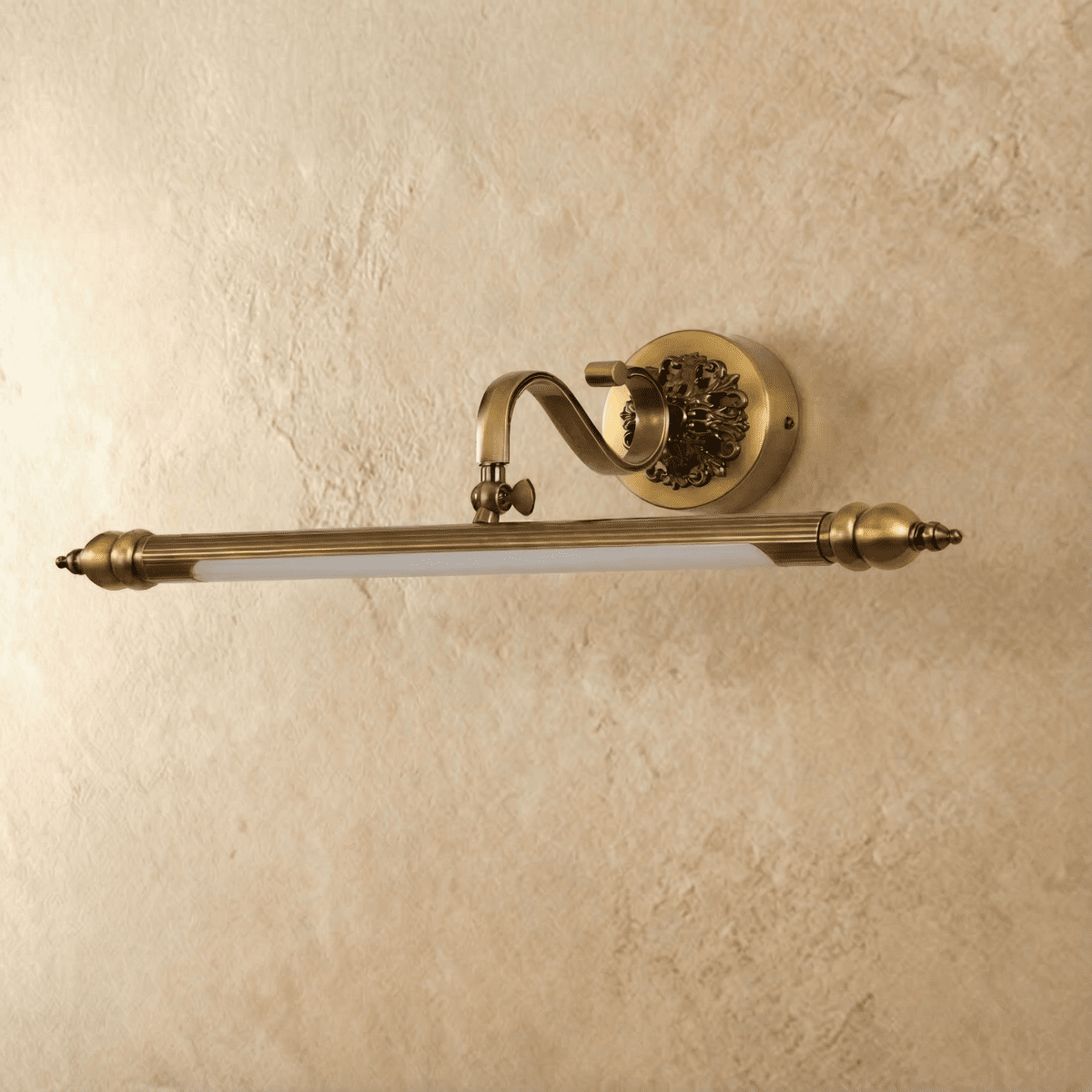 Clérachic antique brass picture-style wall light with a fluted bar, scrolled arm and ornate round backplate, shown unlit on a plaster wall.