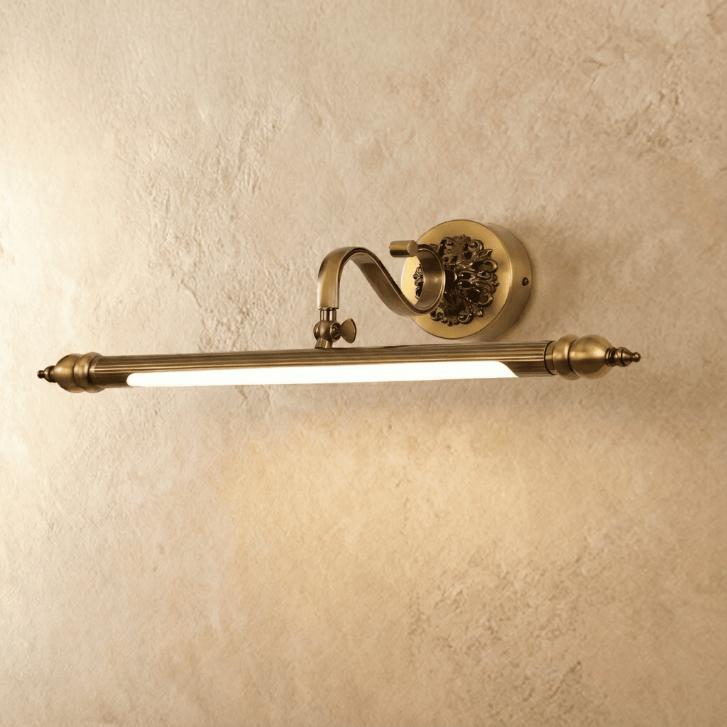 Clérachic antique brass wall light with a fluted bar and scrolled arm, lit with a warm glow against a limewashed plaster wall.