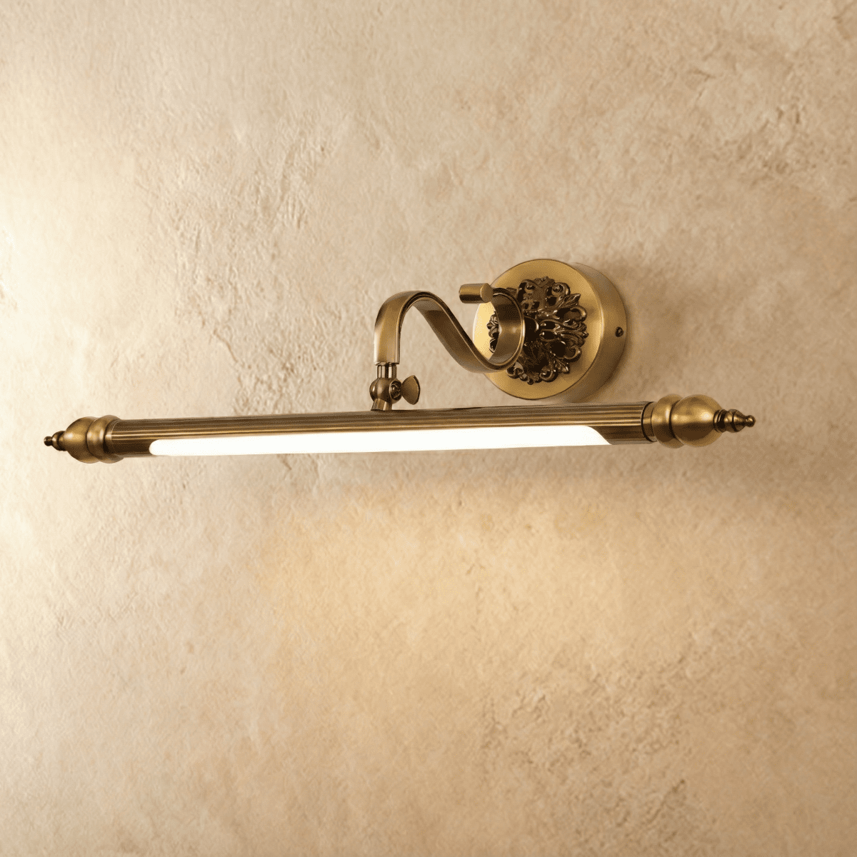 Clérachic antique brass wall light with a fluted bar and scrolled arm, lit with a warm glow against a limewashed plaster wall.
