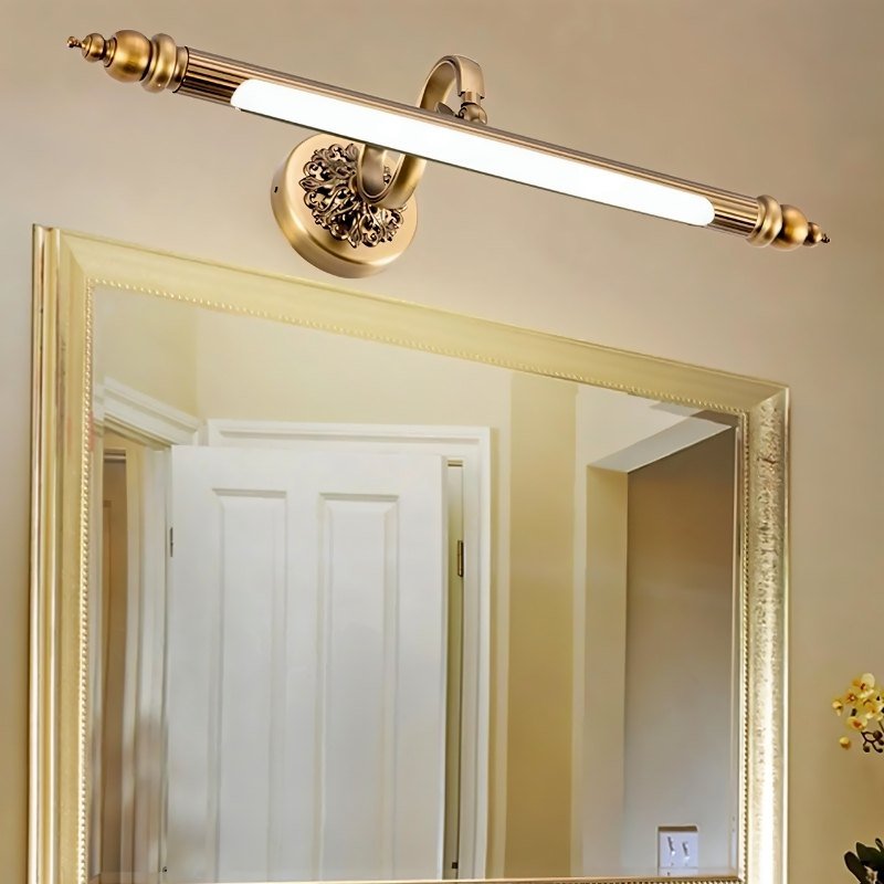 Clérachic antique brass fluted bar light angled over a gold-framed mirror, its ornate round backplate and scrolled arm in view.