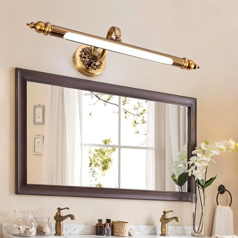 Clérachic antique brass bar light mounted above a dark wood-framed vanity mirror, with brass taps and white flowers beside it.