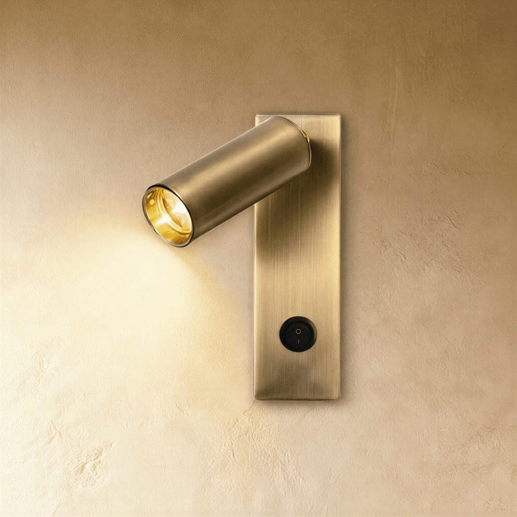 Clérachic slim brushed brass wall reading light with an angled cylindrical head, lit warm, switch at the base of the backplate.