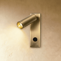 Clérachic slim brushed brass wall reading light with an angled cylindrical head, lit warm, switch at the base of the backplate.