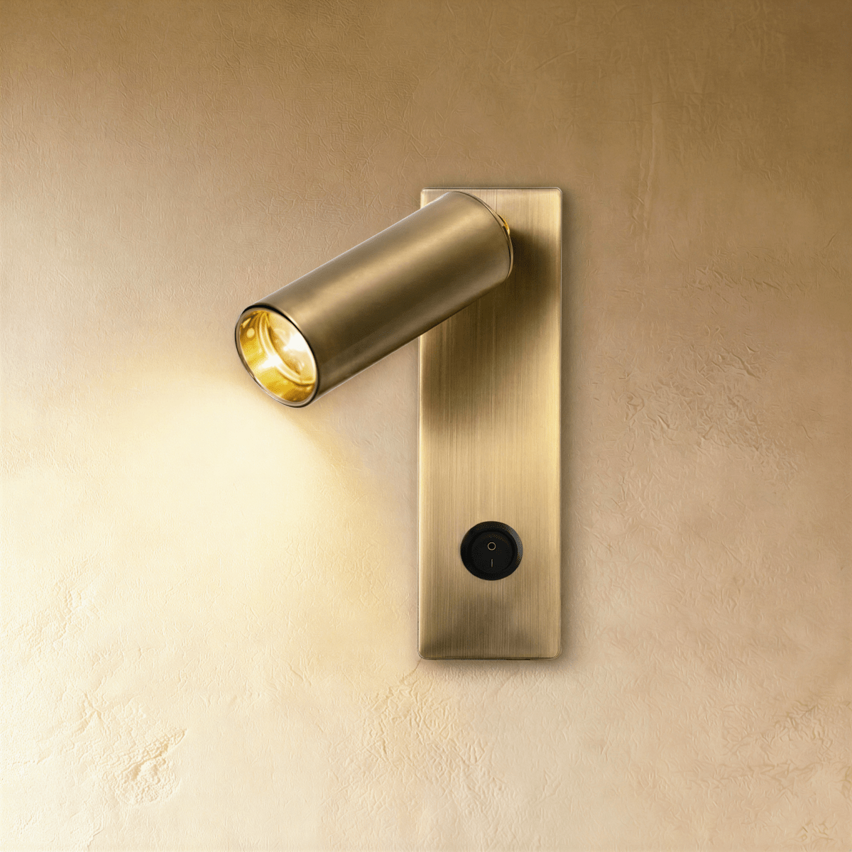 Clérachic slim brushed brass wall reading light with an angled cylindrical head, lit warm, switch at the base of the backplate.
