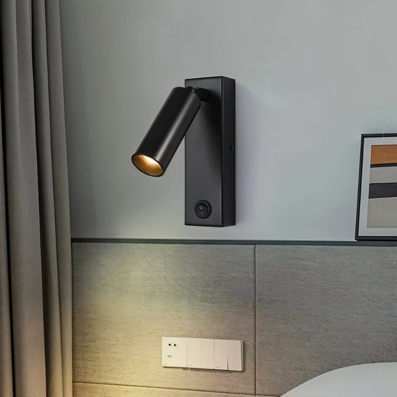Clérachic black wall reading light with a rocker switch, mounted on a pale wall above a grey padded headboard.