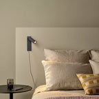 Clérachic dark wall reading light with a USB port, charging a phone by cable above a bedside table and linen bedding.