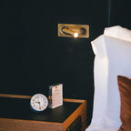 Clérachic brass reading spotlight turned to a bedside table with a clock, against a dark upholstered headboard.