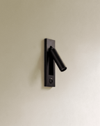 Clérachic black wall reading lamp with a round push button set in the lower plate, cylindrical head angled out on a cream wall.