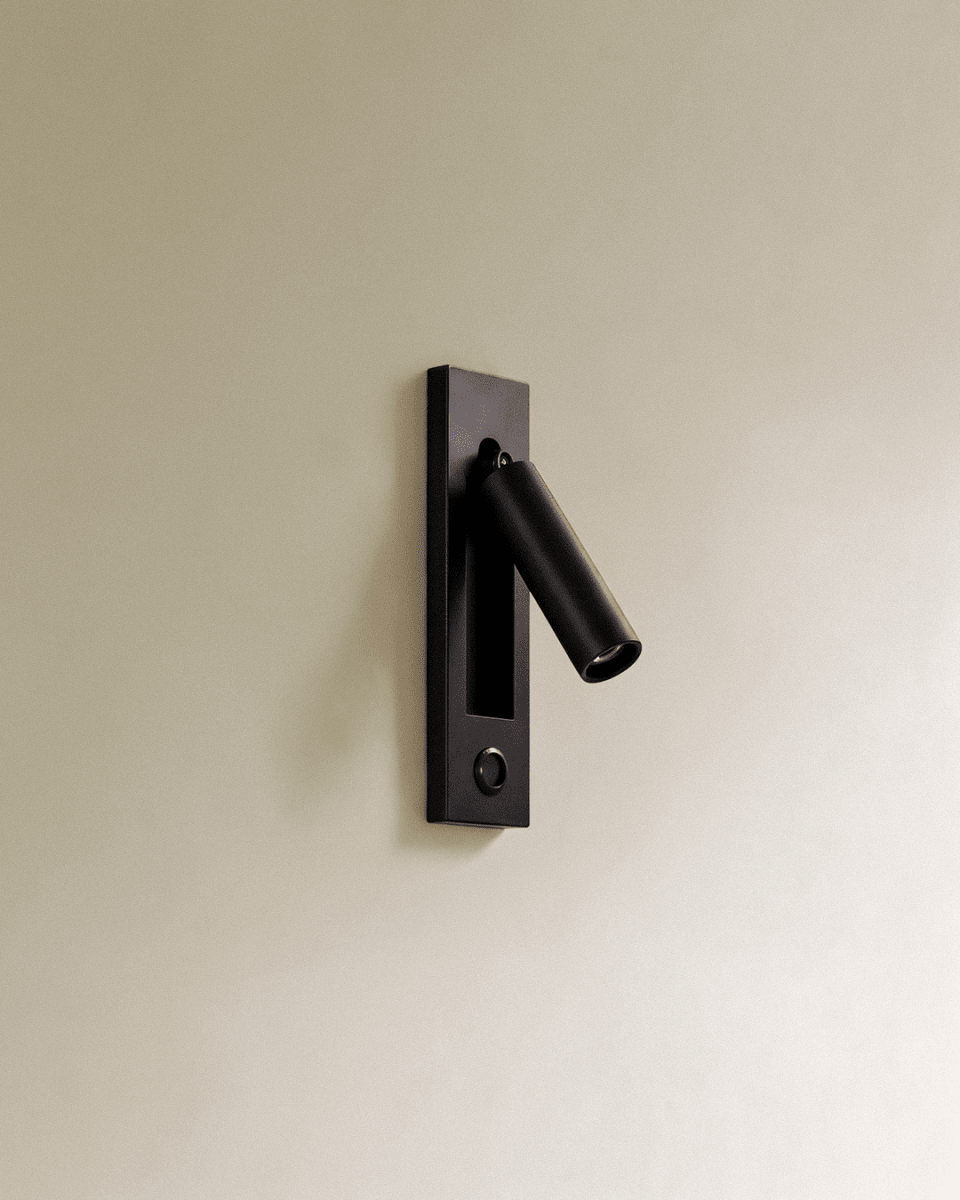 Clérachic black wall reading lamp with a round push button set in the lower plate, cylindrical head angled out on a cream wall.