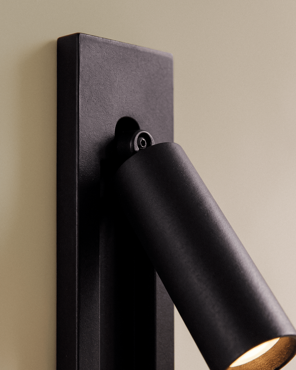 Close view of a Clérachic black wall reading lamp: textured matte plate, small pivot screw and the round lamp head with warm glow.