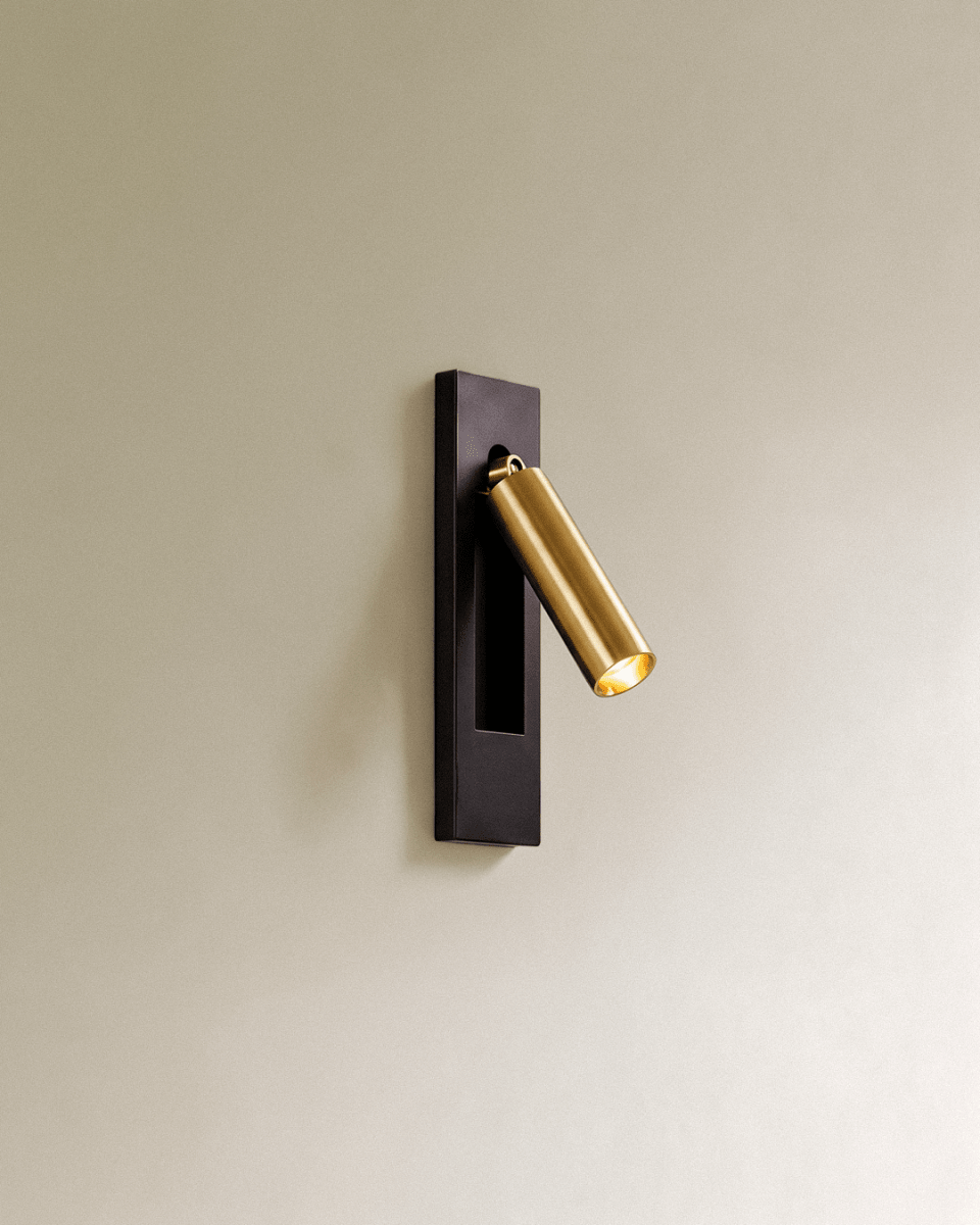 Clérachic wall reading lamp with a black rectangular plate and a brushed gold cylindrical head angled out, glowing warm on a cream wall.