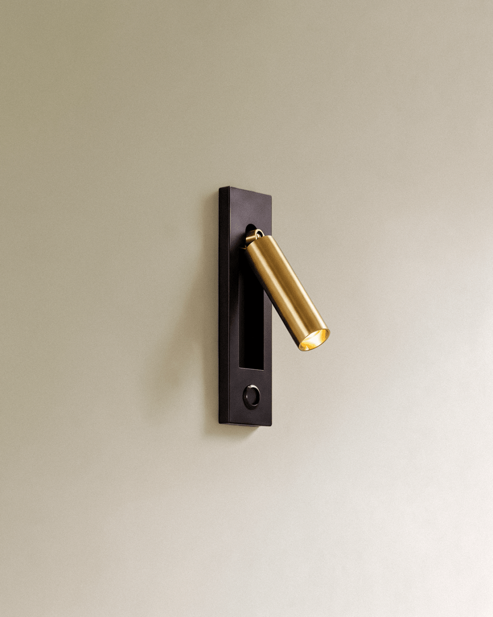 Clérachic black and gold wall reading lamp with a round push button below the recess, gold head angled out on a cream wall.