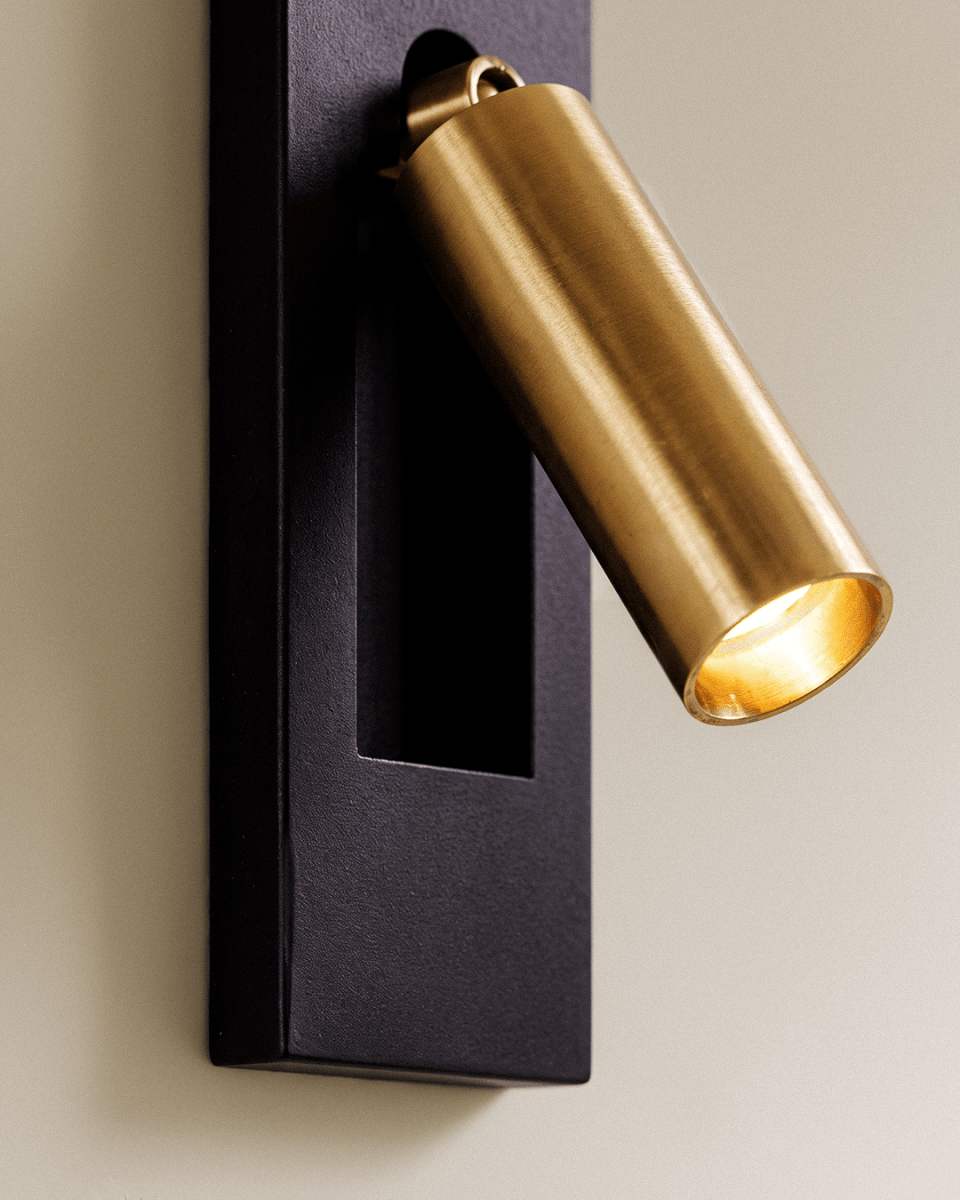 Close view of a Clérachic wall reading lamp: black textured plate with a recessed slot and a brushed gold cylindrical head lit warm.