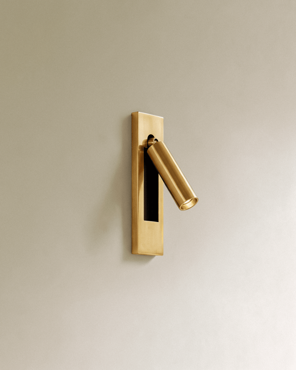 Clérachic all-gold wall reading lamp with a brushed brass-toned plate and cylindrical head angled out on a cream wall.