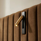 Close view of a Clérachic wall reading lamp with a gold head on a black plate, fixed on a tan channelled velvet headboard.