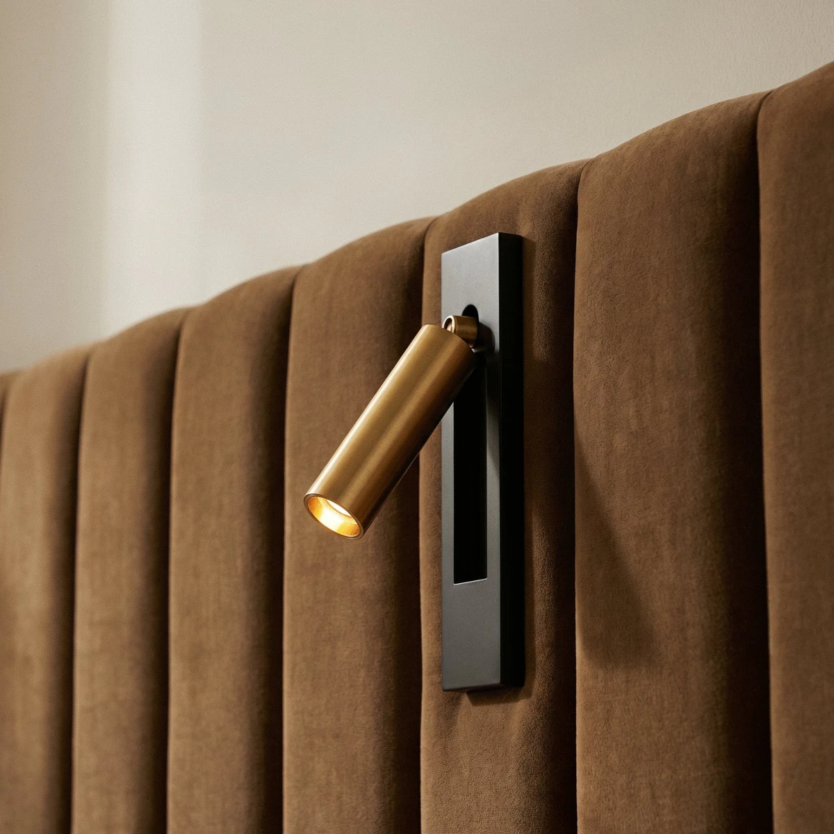 Close view of a Clérachic wall reading lamp with a gold head on a black plate, fixed on a tan channelled velvet headboard.