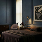 Clérachic wall reading lamp with a gold head on a black plate lighting a bedside corner in a dark blue panelled bedroom.