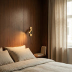 Clérachic all-gold wall reading lamp casting warm light over striped linen bedding against a walnut veneer wall.