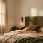 Clérachic black wall reading lamp angled over a walnut floating nightstand beside an olive boucle headboard and linen bedding.