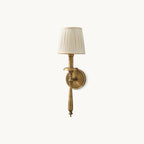 Antique brass wall light with a beige pleated fabric shade, a slim turned stem and a round backplate, shown as a Clérachic sconce on a plain backdrop.
