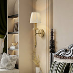 Antique brass Clérachic wall sconce with a pleated shade lit beside a carved dark bedpost and a recessed shelf with a green curtain.