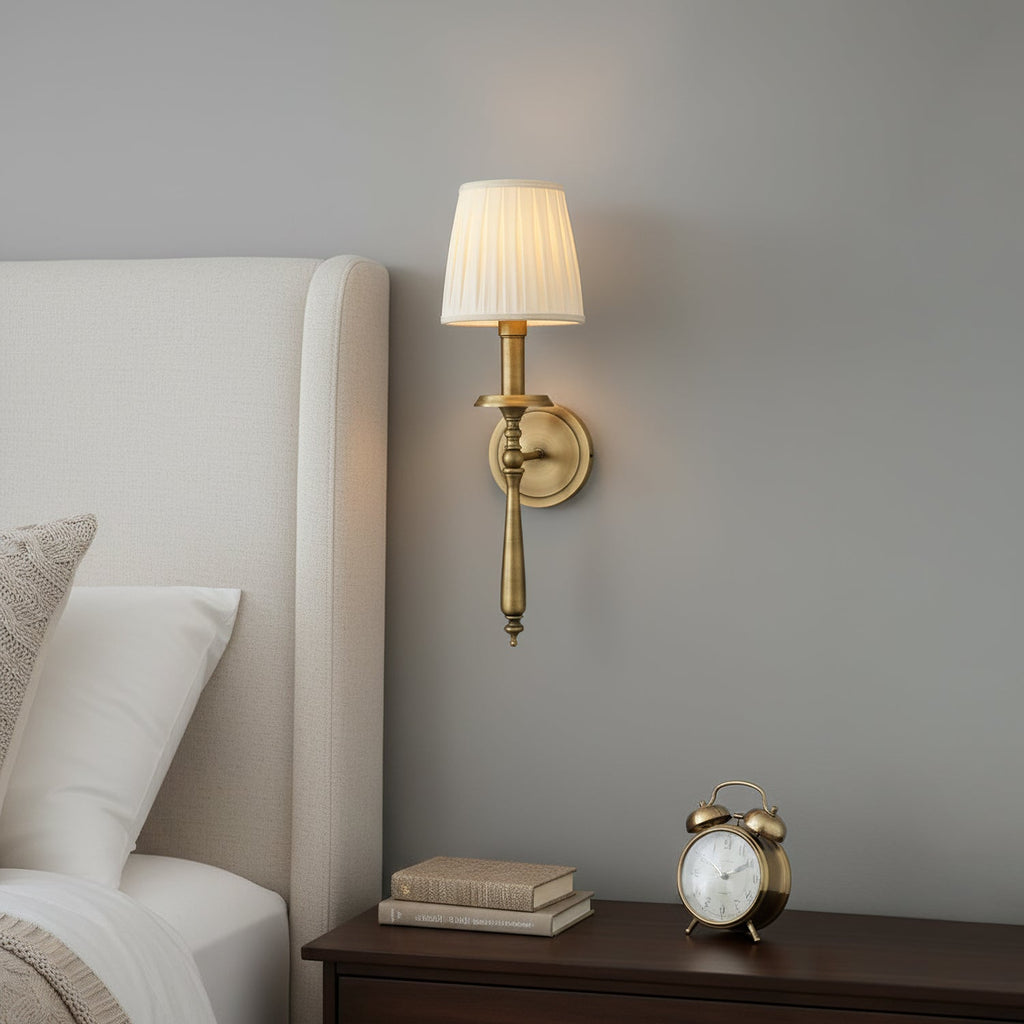 A Clérachic brass wall light with a pleated beige shade, mounted on a grey wall above a bedside table with a small alarm clock.