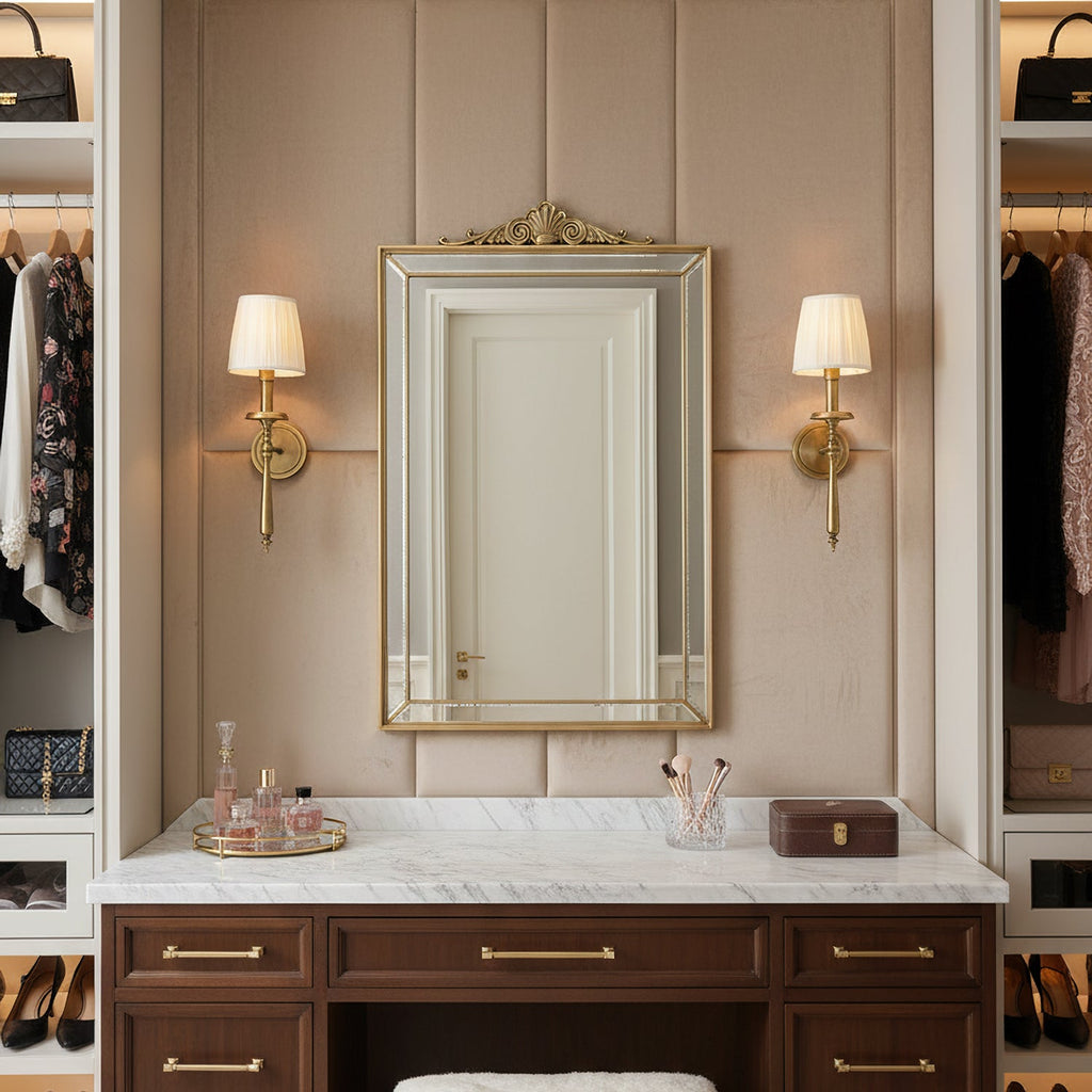 Two Clérachic brass sconces with pleated shades flank an arch-topped mirror above a marble-topped dressing table in a dressing room.