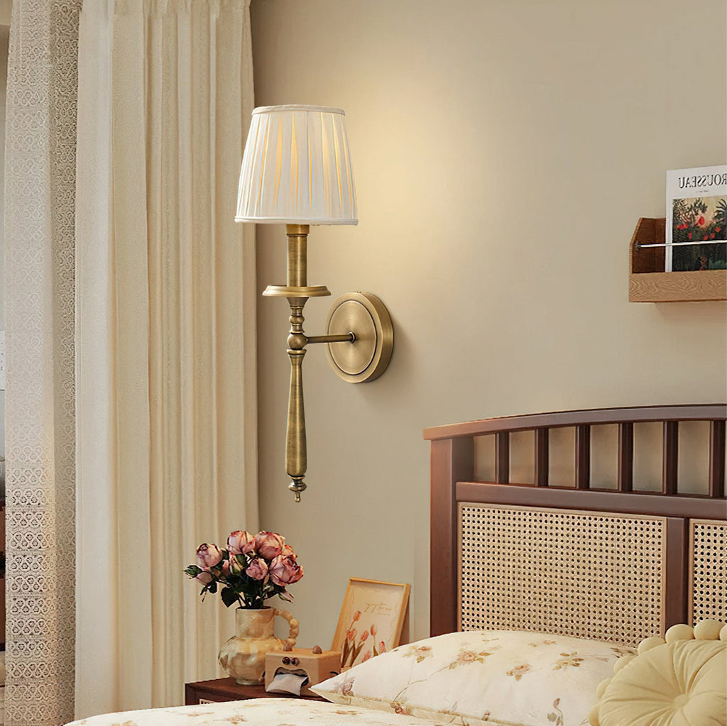 Lit Clérachic brass wall sconce with a pleated shade above a bedside table with pink roses and a cane-headed bed.