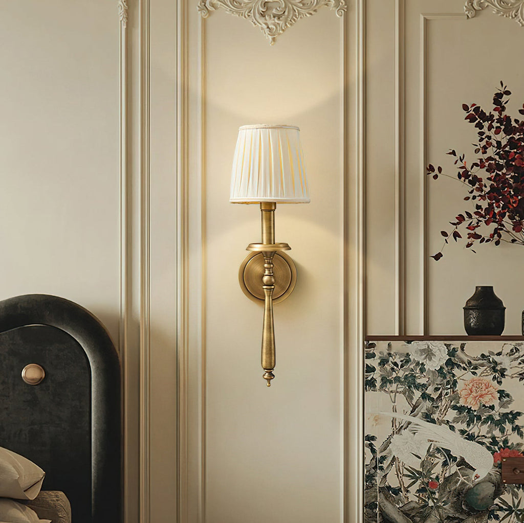 A Clérachic wall light in antique brass with a pleated shade glows on a cream panelled wall, beside a painted floral screen.