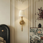 A Clérachic wall light in antique brass with a pleated shade glows on a cream panelled wall, beside a painted floral screen.