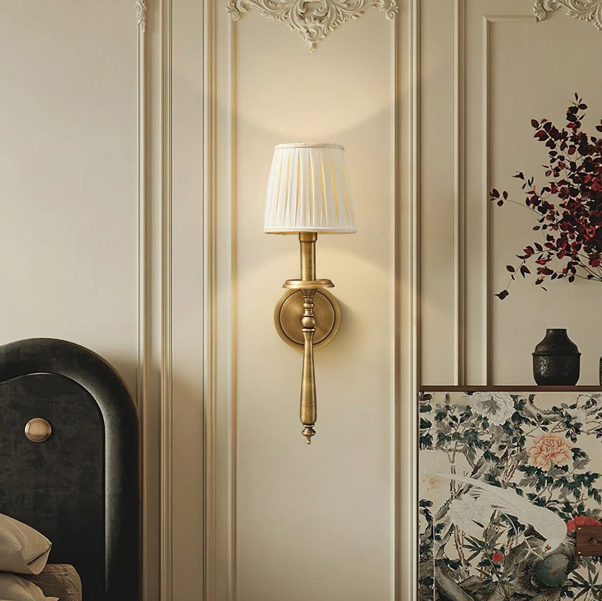 A Clérachic wall light in antique brass with a pleated shade glows on a cream panelled wall, beside a painted floral screen.