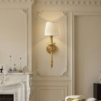 Brass Clérachic wall sconce with a pleated shade glowing against ornate cream wall mouldings, above a carved marble fireplace.