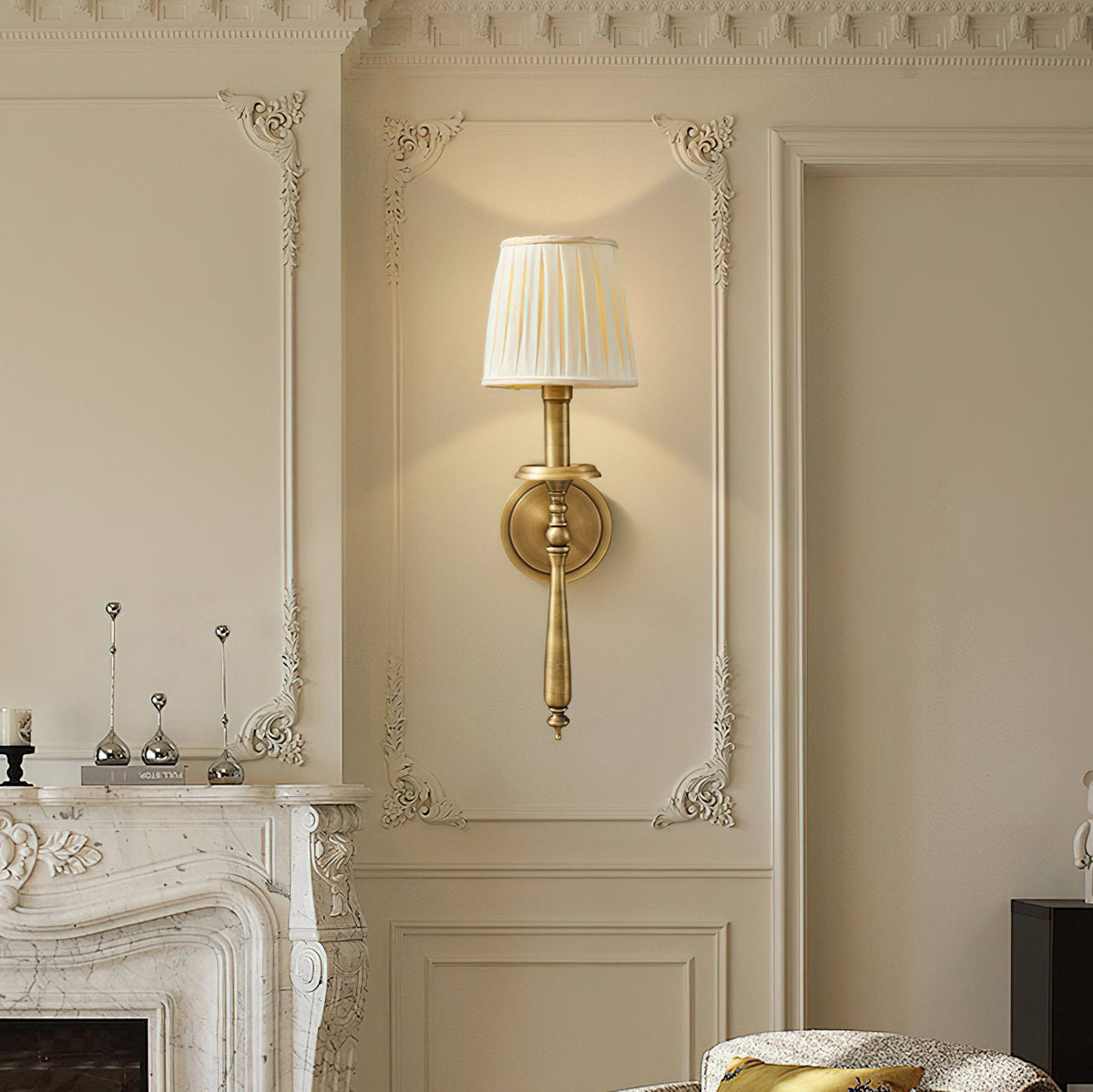 Brass Clérachic wall sconce with a pleated shade glowing against ornate cream wall mouldings, above a carved marble fireplace.