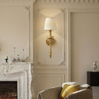 Clérachic wall light with a pleated beige shade on a cream moulded wall, above a marble fireplace and a speckled armchair.