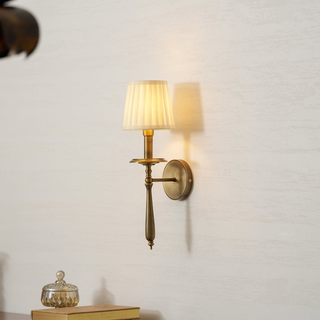 Lit Clérachic wall sconce in antique brass with a pleated shade, casting a soft shadow on a pale textured wall.