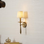 Lit Clérachic wall sconce in antique brass with a pleated shade, casting a soft shadow on a pale textured wall.