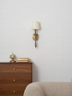 Brass Clérachic wall light mounted above a walnut chest of drawers and a cream sofa, with a glass trinket jar and books beside it.