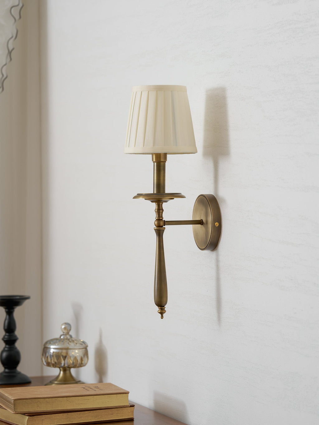 Side view of a Clérachic wall sconce showing its brass arm, round backplate and pleated shade, above a stack of books.