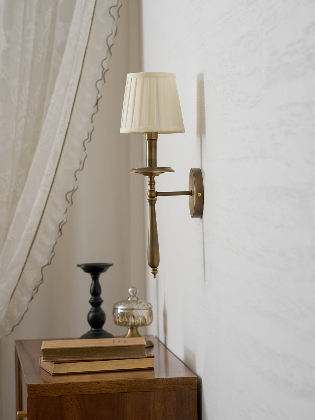 A Clérachic brass wall light with a pleated shade beside a sheer curtain, above a wooden side table with a black candlestick.