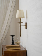 A Clérachic brass wall light with a pleated shade beside a sheer curtain, above a wooden side table with a black candlestick.