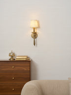 Lit Clérachic wall sconce on a pale wall above a walnut chest of drawers, with a glass jar and a cream sofa arm.