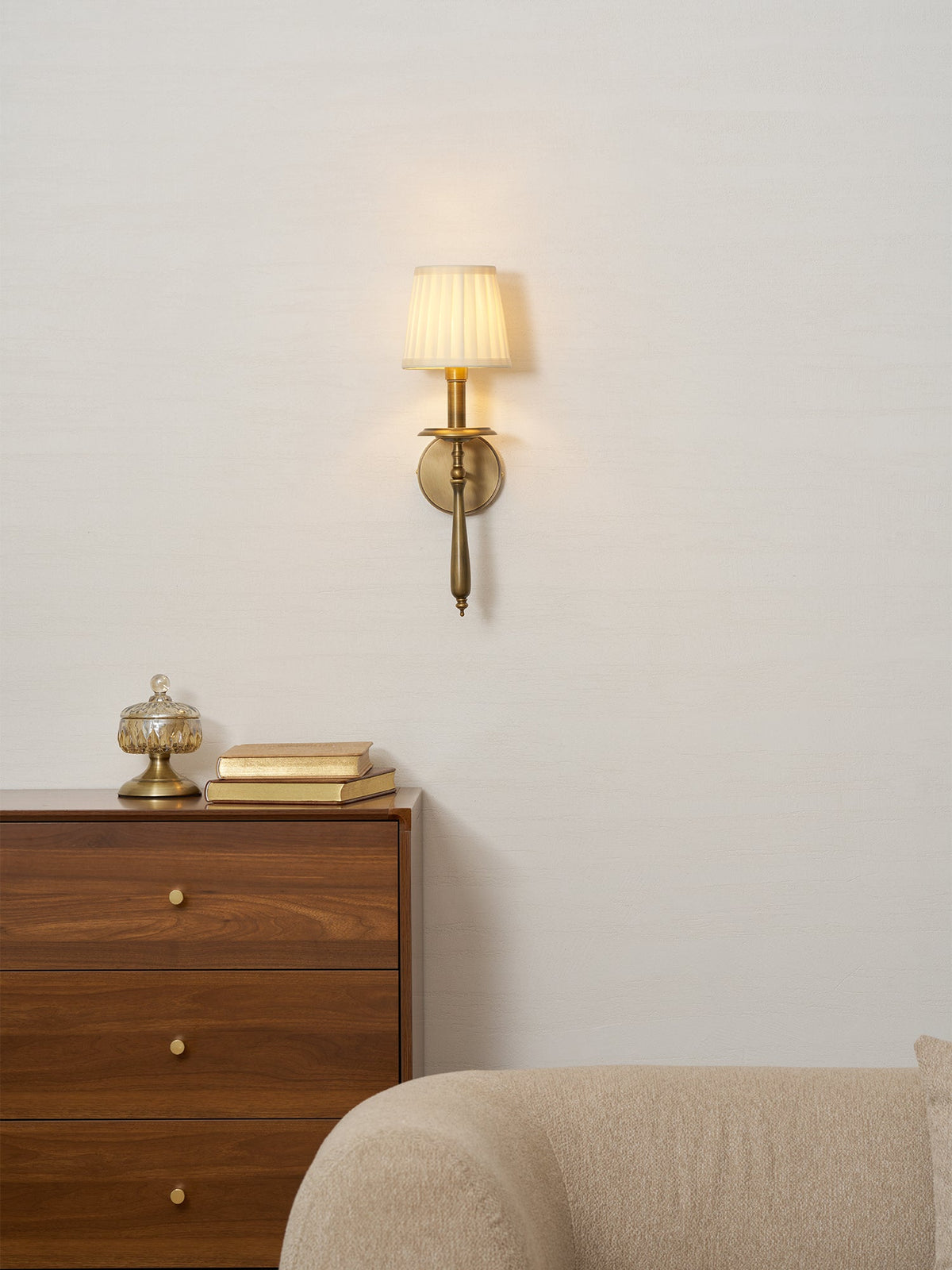 Lit Clérachic wall sconce on a pale wall above a walnut chest of drawers, with a glass jar and a cream sofa arm.
