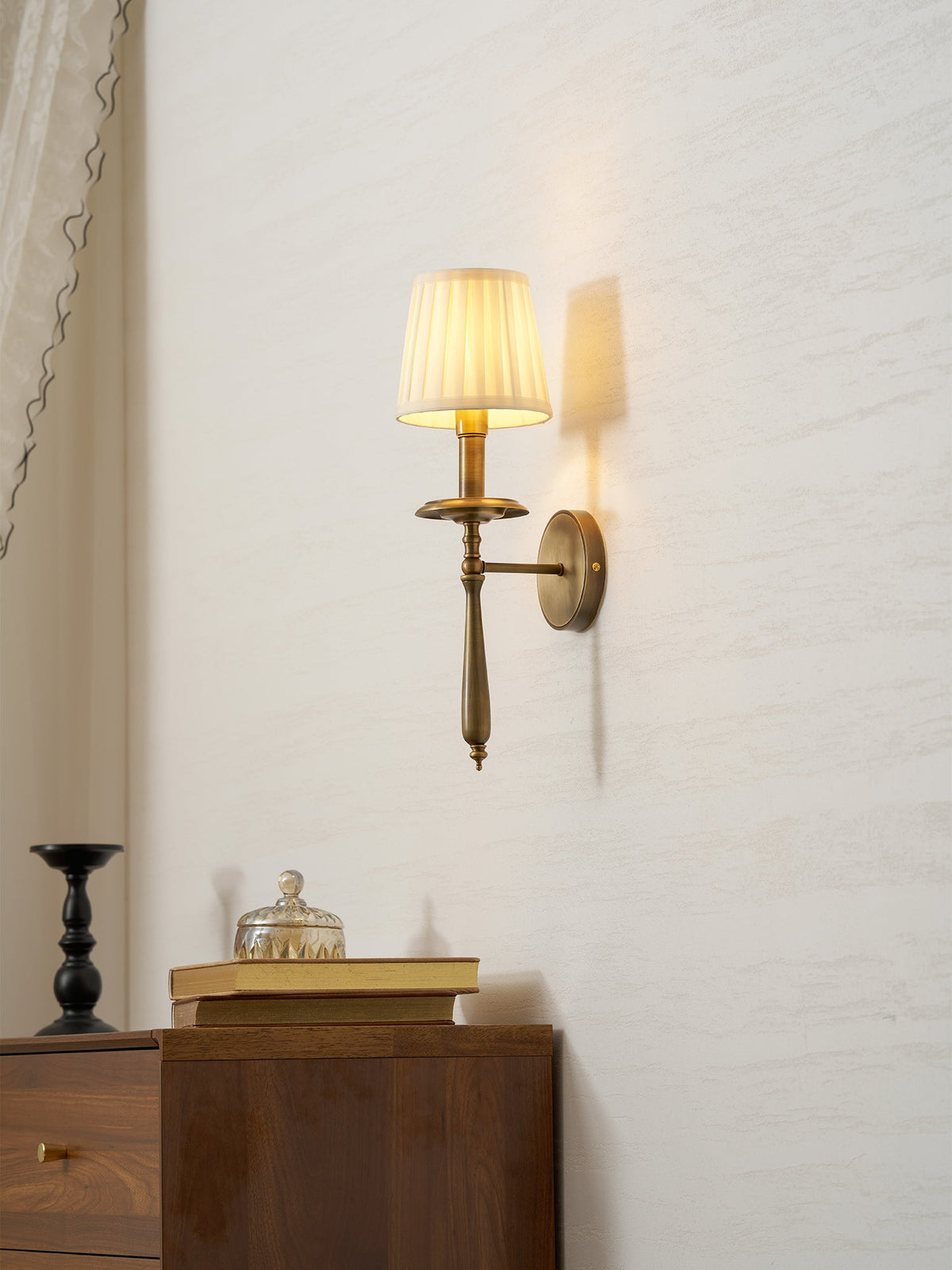 A Clérachic wall sconce glows warm beside a sheer curtain, above a dark candlestick, books and a glass jar.