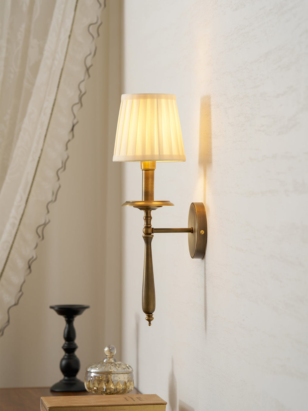 Brass Clérachic wall light with a pleated shade lit softly, set beside a draped curtain, with a black candlestick below.