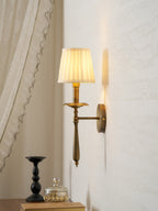Brass Clérachic wall light with a pleated shade lit softly, set beside a draped curtain, with a black candlestick below.