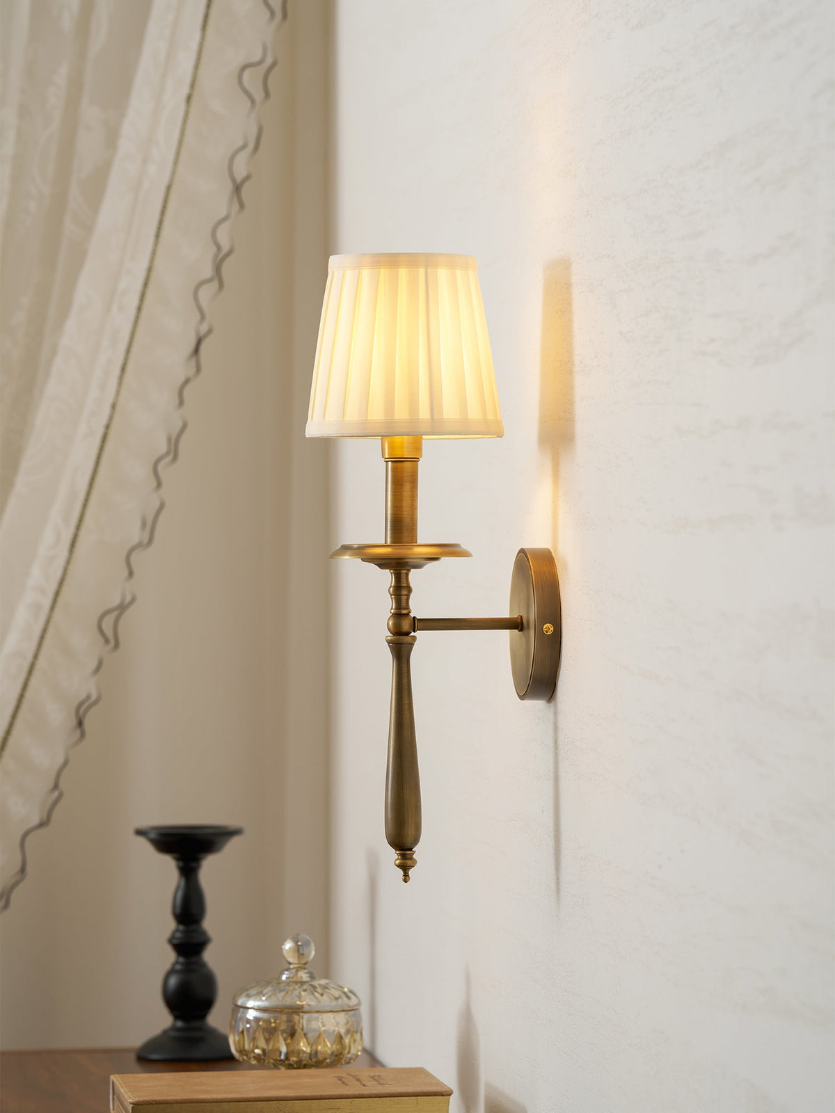 Brass Clérachic wall light with a pleated shade lit softly, set beside a draped curtain, with a black candlestick below.