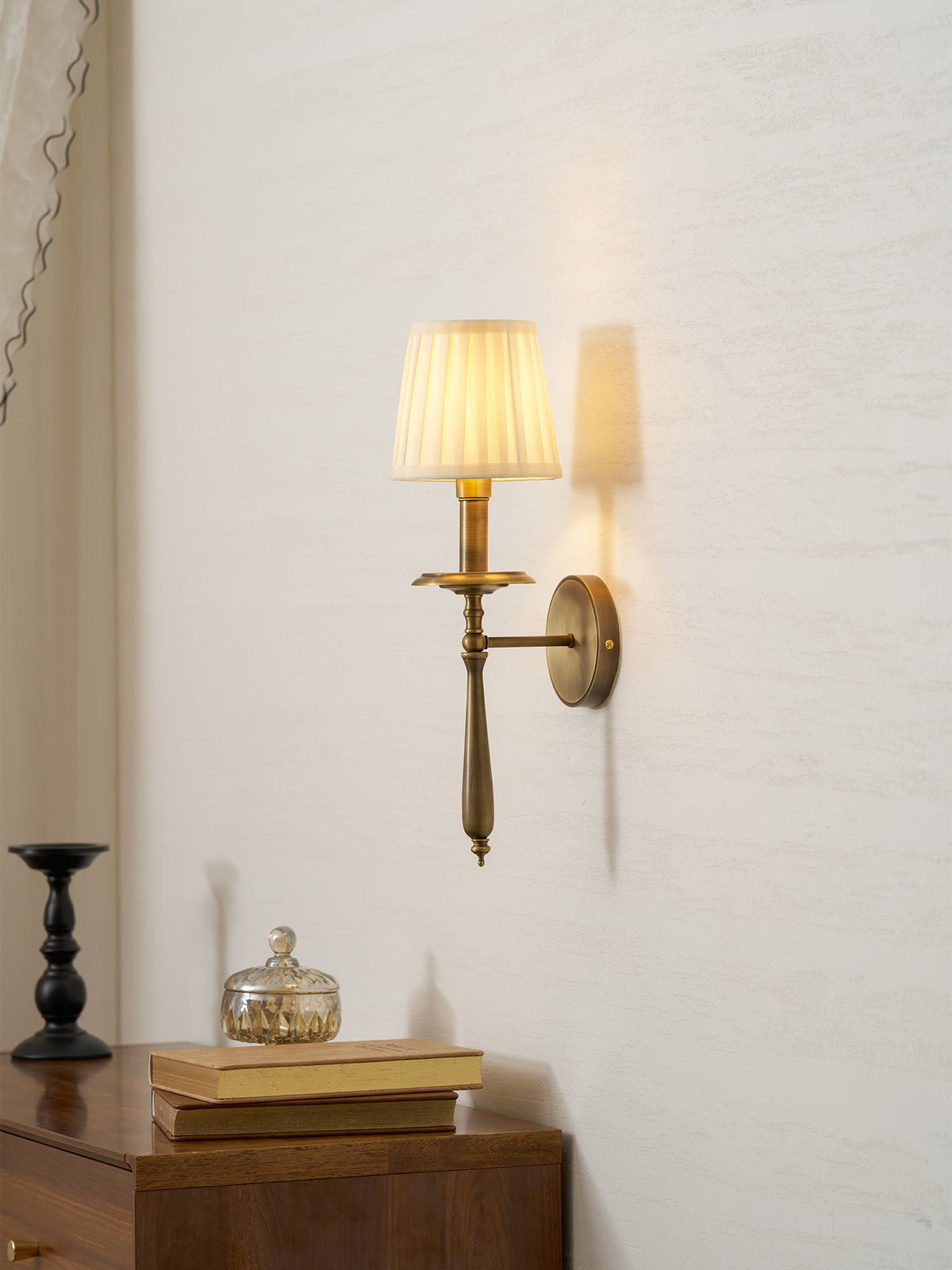 A Clérachic wall light with a pleated shade glowing above a wooden side table with books and a glass jar.